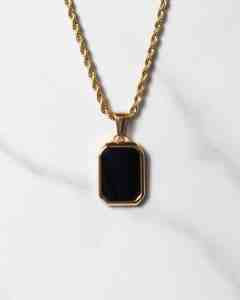 Ett halsband med repkedja i guld och ett rektangulärt hänge i svart onyx med konturer i guld, på en vit marmoryta.