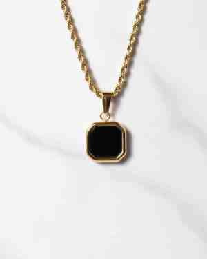 Halsband med repkedja i guld och ett åttkantigt hänge i svart onyx som omges av en guldram, elegant placerat på en vit marmorbakgrund.