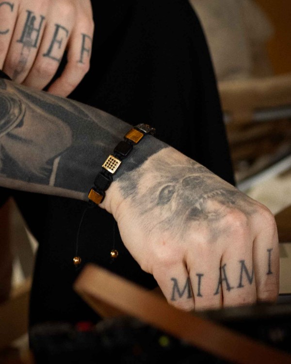 En närbild av tatuerade händer, en med CHEF och den andra med MIAMI på knogarna, visar ett Flatbead Marrakech-armband på handleden och en hundansiktstatuering på underarmen.