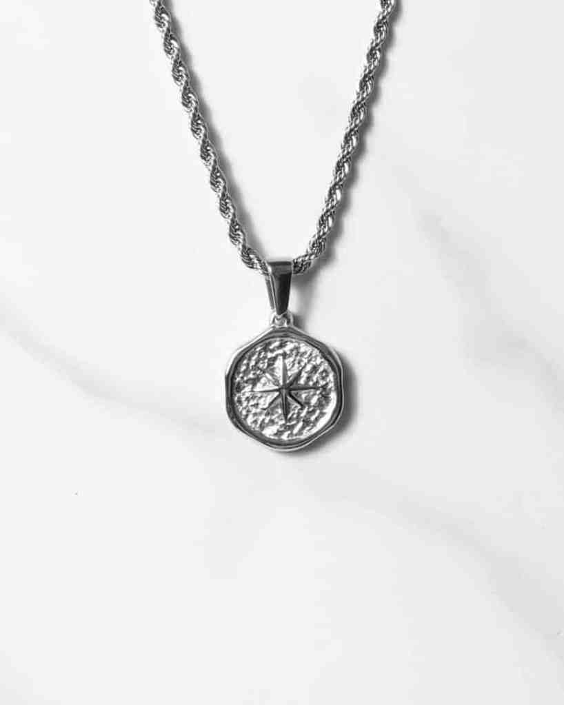 Ett Étoile Sauvage Silverhängehalsband med en texturerad rund medaljong med en kompassdesign, som visas på en tvinnad kedja mot en vit marmorbakgrund.