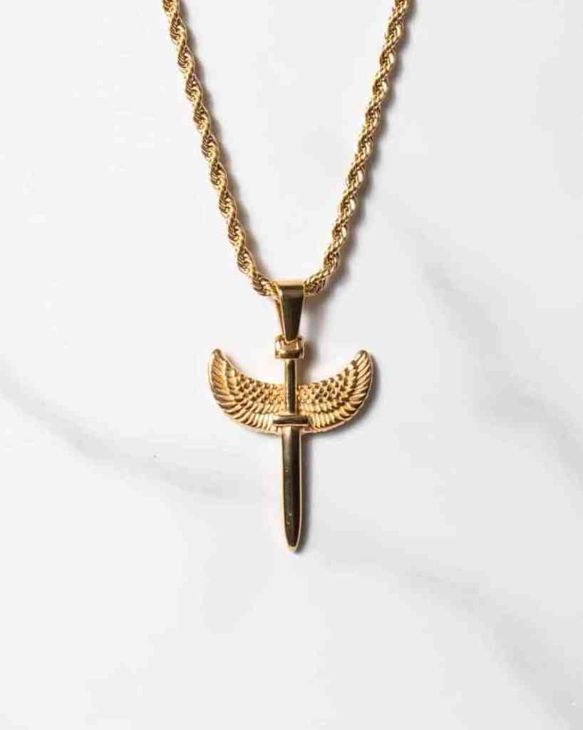 Ett guldhalsband med repkedja och ett Sword Guld-hänge i form av ett vertikalt svärd med utsträckta vingliknande detaljer på vardera sidan, mot en bakgrund av vit marmor.